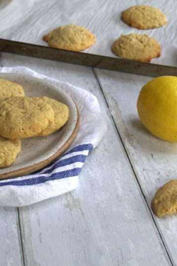 A plate of gluten free lemon cookies and a broken cookie in the forefront of the picture - a lemon and a baking sheet with additional cookies are in the backgrounf