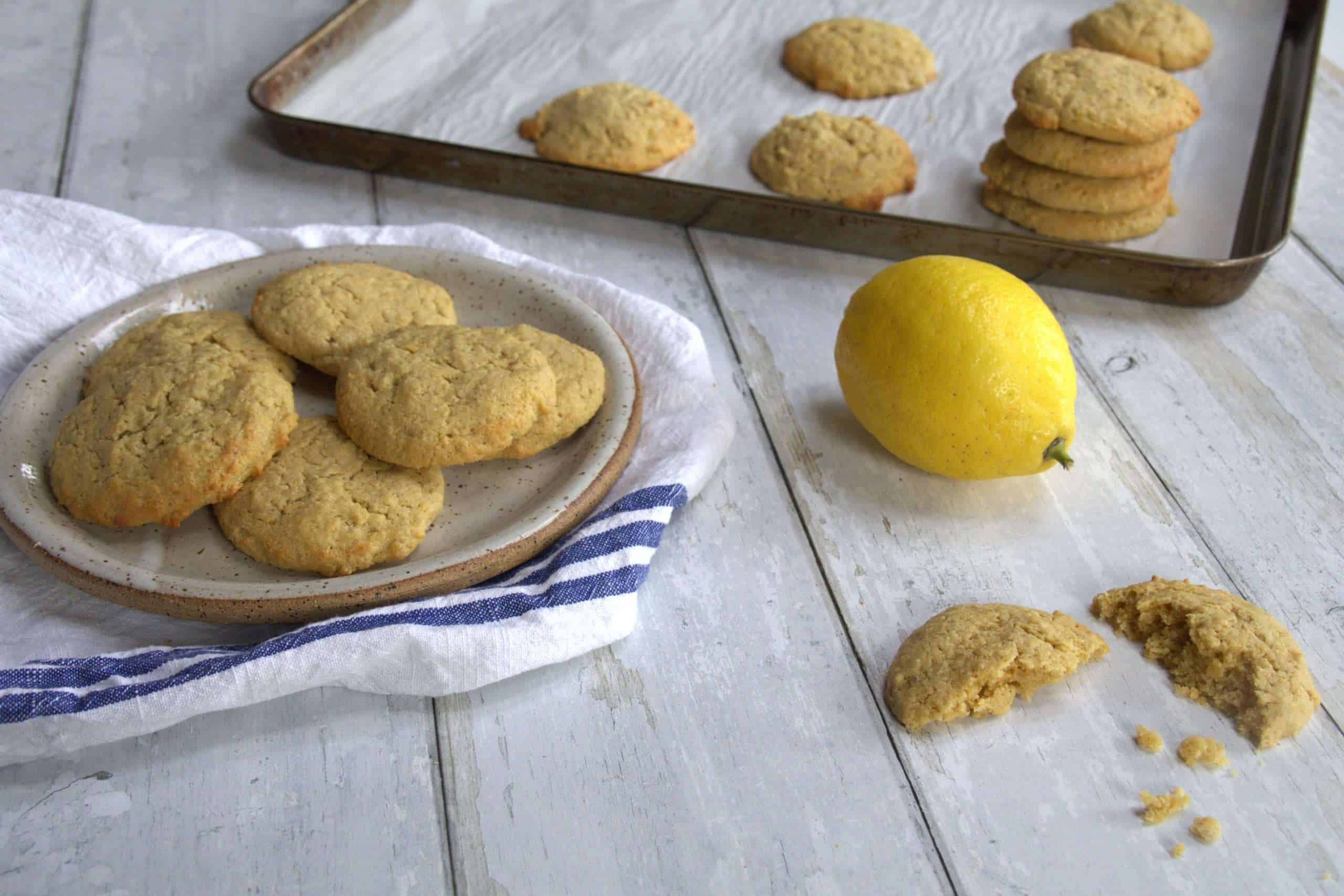 A plate of gluten free lemon cookies and a broken cookie in the forefront of the picture - a lemon and a baking sheet with additional cookies are in the backgrounf