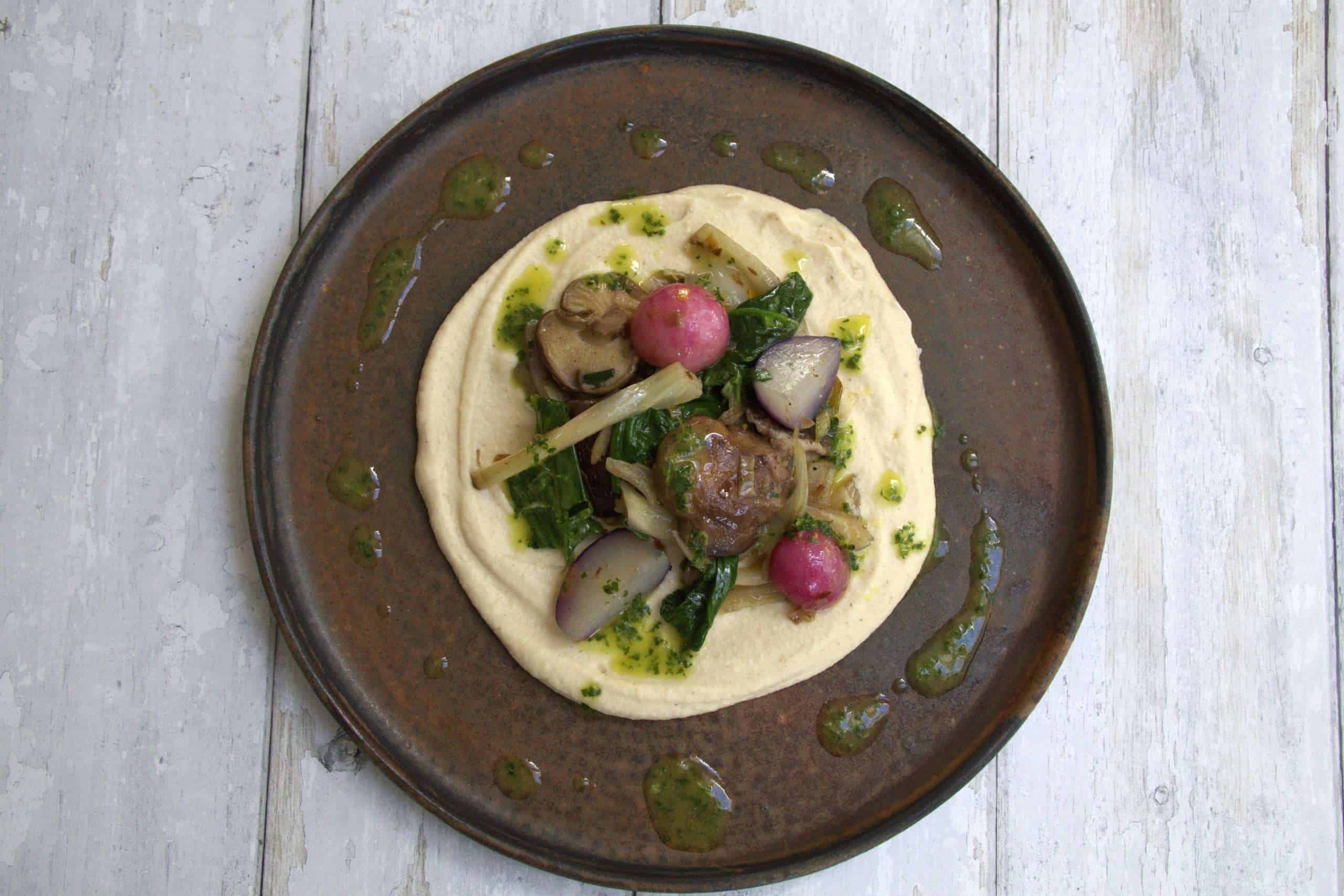 A beautiful dark colored plate with the assembled spring vegetables and white bean cauliflower purée - a layer of white bean and cauliflower puree topped with sauteed vegetables and drizzled with lemon and herb oil