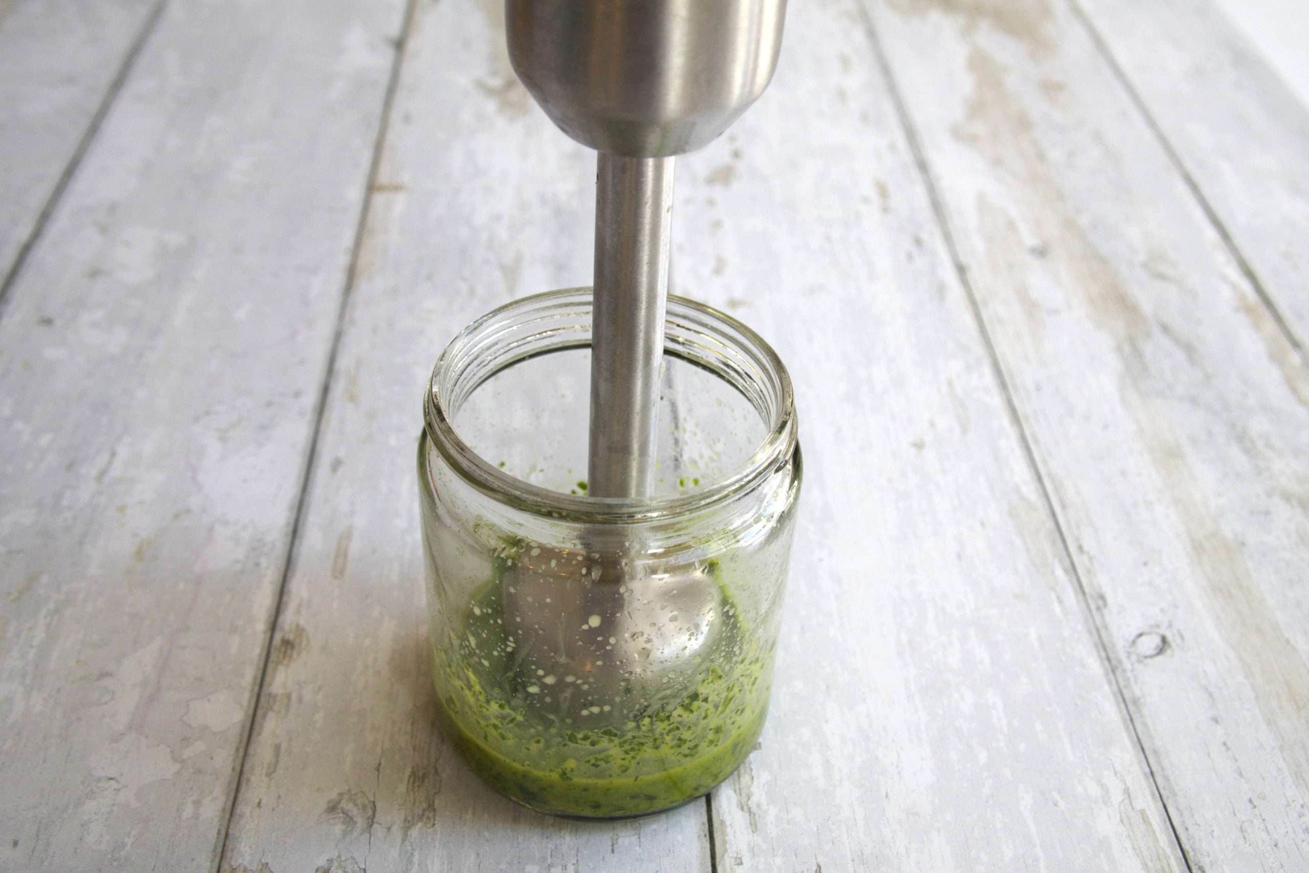 A small glass container holding olive oil, parsley and lemon zest with an immersion blender ready to blend