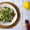 A bowl with roasted spring vegetables, including radishes, spring onions and snap peas, drizzled with an herby sauce, is sitting on top of a wooden board - a whole lemon and fresh radishes are displayed along with the bowl