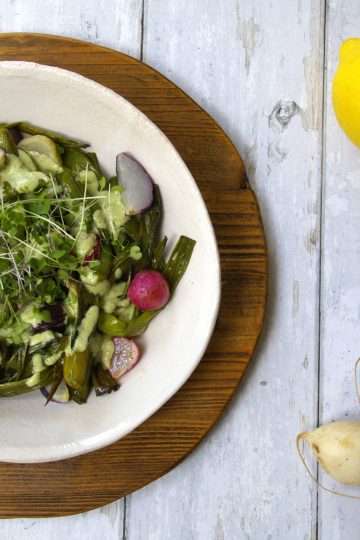A bowl with roasted spring vegetables, including radishes, spring onions and snap peas, drizzled with an herby sauce, is sitting on top of a wooden board - a whole lemon and fresh radishes are displayed along with the bowl