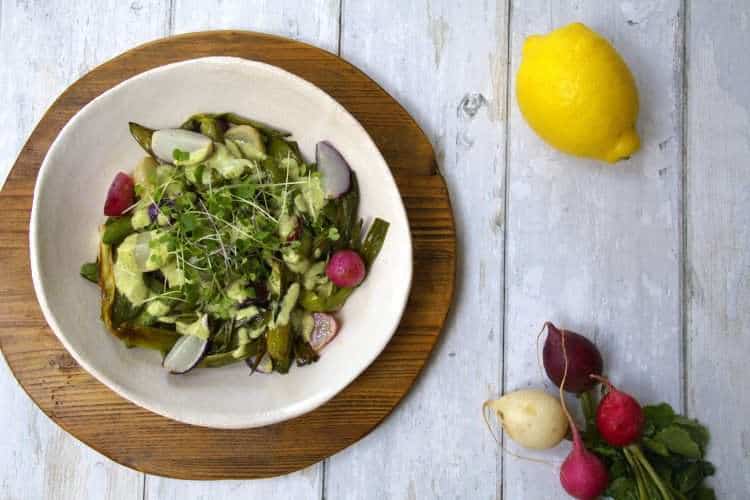 A bowl with roasted spring vegetables, including radishes, spring onions and snap peas, drizzled with an herby sauce, is sitting on top of a wooden board - a whole lemon and fresh radishes are displayed along with the bowl