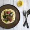 a dark colored plate topped with creamy cauliflower and white bean puree and sauteed springe vegetables - the outer edge is drizzled with herb and lemon oil and there is half a squeezed lemon on the side along with a dark colored spoon and fork