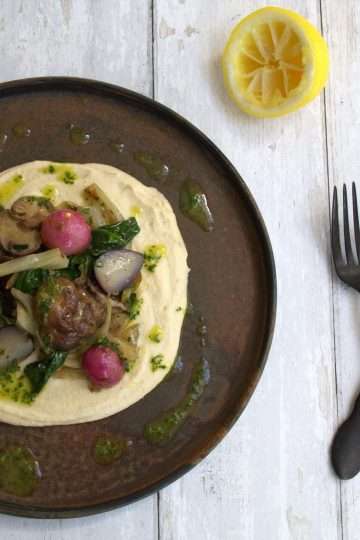 a dark colored plate topped with creamy cauliflower and white bean puree and sauteed springe vegetables - the outer edge is drizzled with herb and lemon oil and there is half a squeezed lemon on the side along with a dark colored spoon and fork