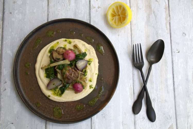 a dark colored plate topped with creamy cauliflower and white bean puree and sauteed springe vegetables - the outer edge is drizzled with herb and lemon oil and there is half a squeezed lemon on the side along with a dark colored spoon and fork