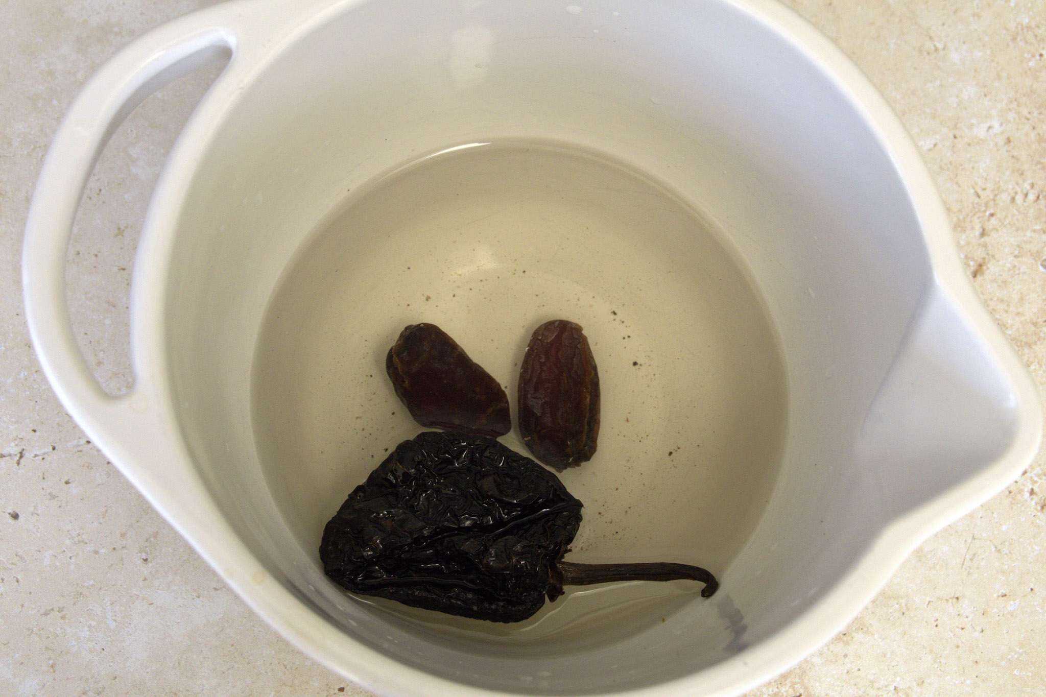 Two dates and one ancho chili soaking in water in a white bowl