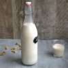 A bottle of nut milk on a grey surface with a wooden background - a small glass of nut milk is next to the bottle and some walnuts are scattered on the ground
