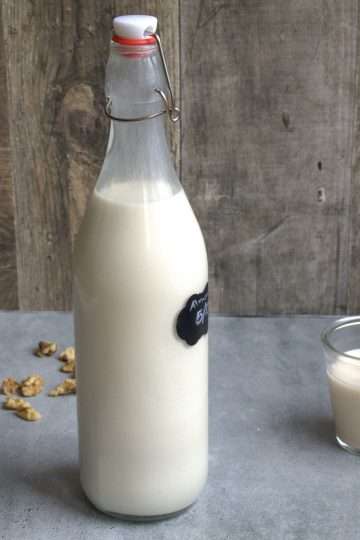 A bottle of nut milk on a grey surface with a wooden background - a small glass of nut milk is next to the bottle and some walnuts are scattered on the ground