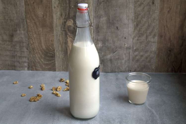 A bottle of nut milk on a grey surface with a wooden background - a small glass of nut milk is next to the bottle and some walnuts are scattered on the ground