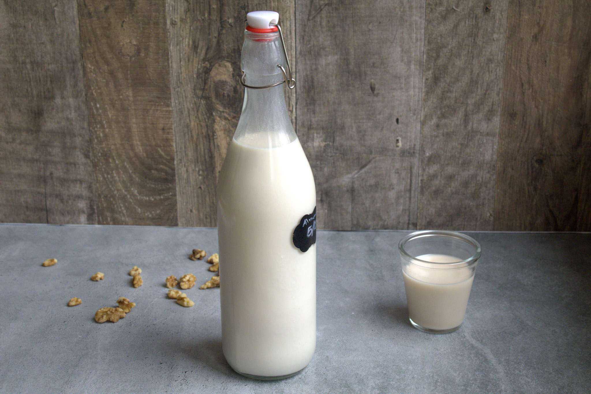A bottle of nut milk on a grey surface with a wooden background - a small glass of nut milk is next to the bottle and some walnuts are scattered on the ground