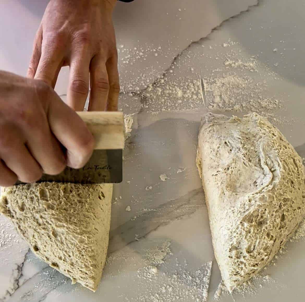 Two hands can be seen cutting a batch of overnight fermented sourdough with a bench scraper