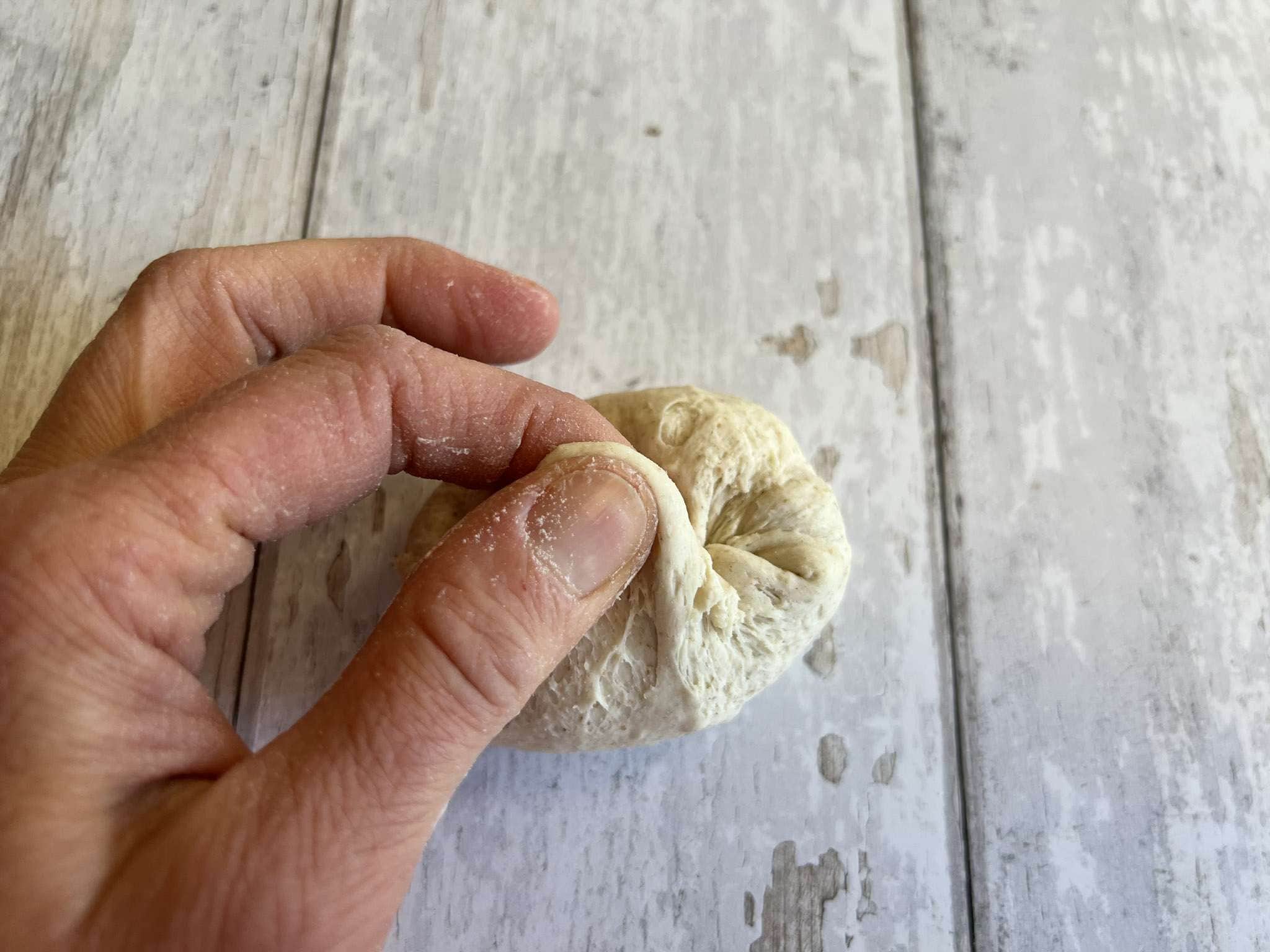a hand can be seen shaping and pinching a sourdough ball into pretzel roll shape