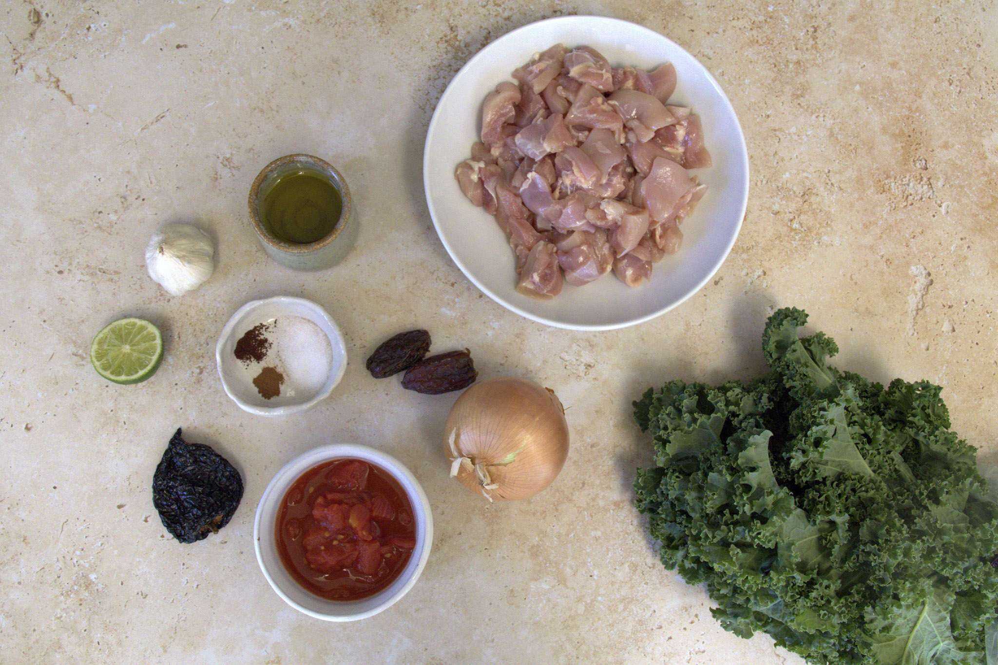 All ingredients for adobo-inspired chicken and kale tacos displayed, including chicken, kale, onion, dates, diced tomatoes, olive oil, fine sea salt, ground cinnamon and cloves, garlic, ancho chili and lime