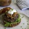 A white pottery plate with bison burger, made of a pretzel roll half topped with arugula, a bison burger patty, a spoonful of rhubarb chutney and a dollop of goat cheese cream - the plate is sprinkled with chives and there is a grey and white striped napkin in the back along with a jar of rhubarb chutney