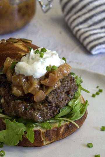 A white pottery plate with bison burger, made of a pretzel roll half topped with arugula, a bison burger patty, a spoonful of rhubarb chutney and a dollop of goat cheese cream - the plate is sprinkled with chives and there is a grey and white striped napkin in the back along with a jar of rhubarb chutney