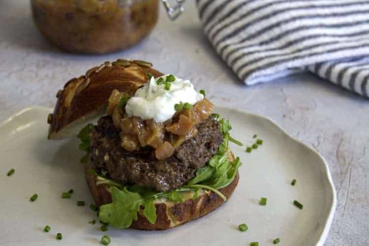 A white pottery plate with bison burger, made of a pretzel roll half topped with arugula, a bison burger patty, a spoonful of rhubarb chutney and a dollop of goat cheese cream - the plate is sprinkled with chives and there is a grey and white striped napkin in the back along with a jar of rhubarb chutney