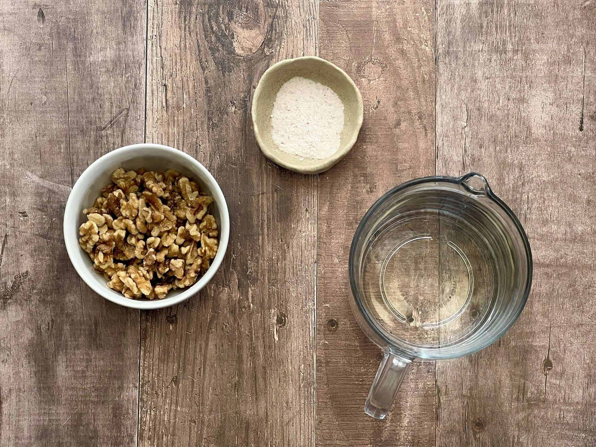 ingredients for nut milk are displayed on a wooden surface, including a bowl of walnuts, a pitcher with water and a small bowl with fine sea salt