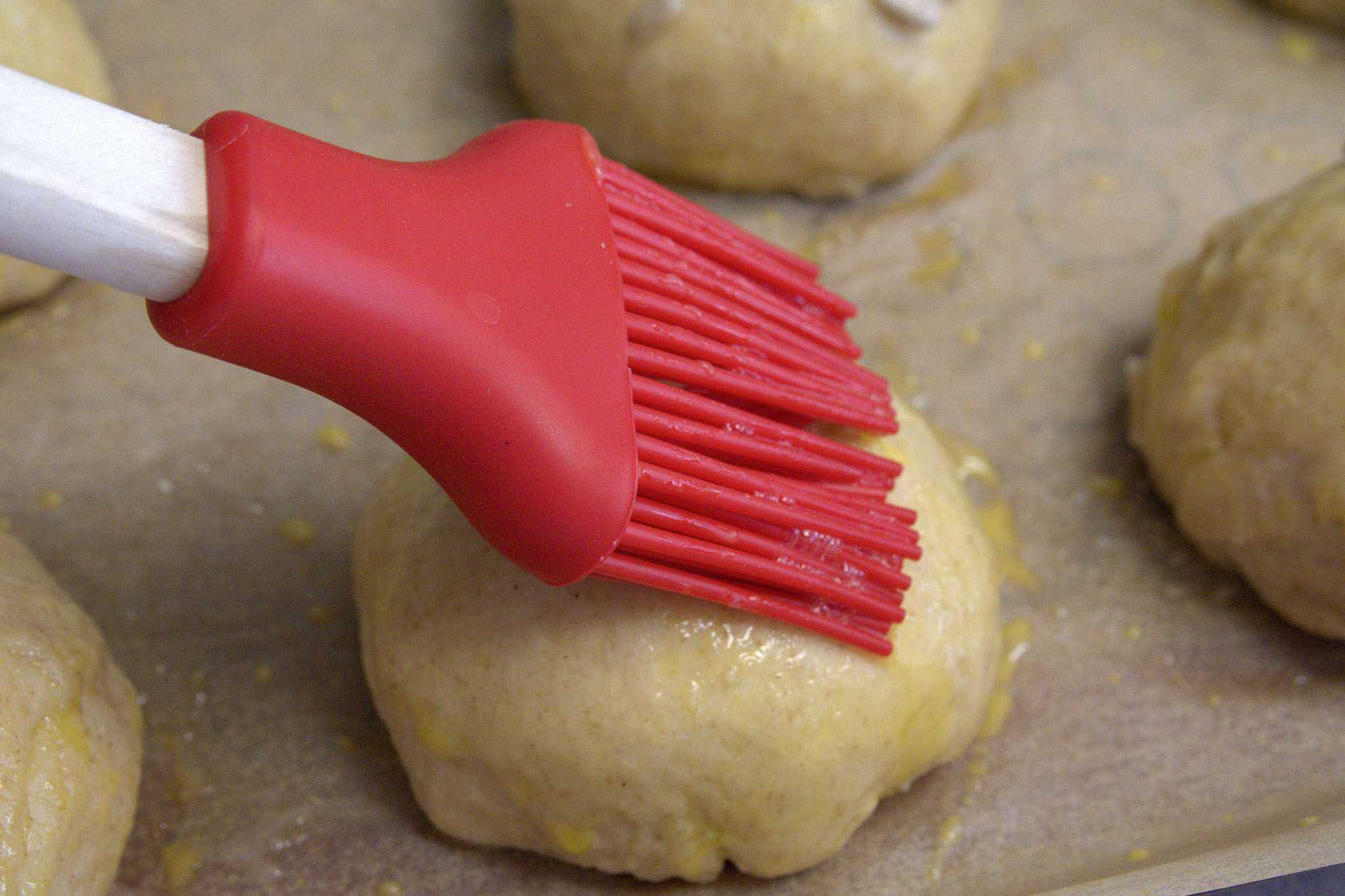 A previously boiled sourdough pretzel roll on a parchment lined baking sheet is being brushed with egg wash with a red silicone brush
