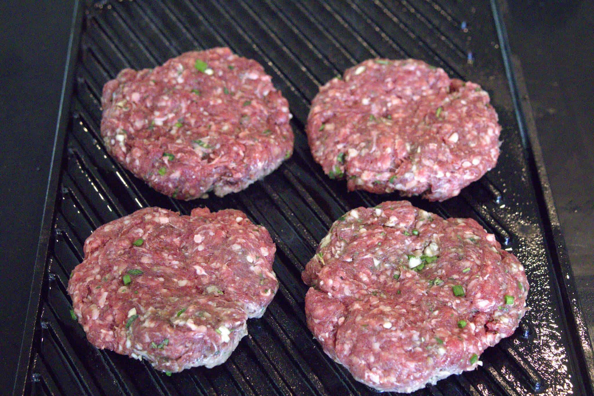 4 bison burger patties cooking on a grill pan - they are showing the beginnings of being cooked on the bottom, but are still raw on top