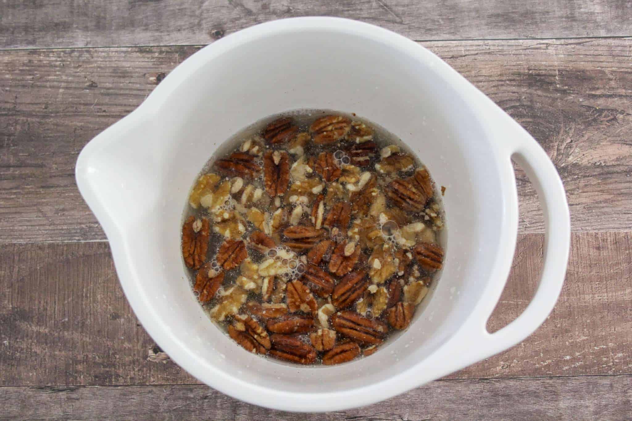 a white bowl resting on a wooden surface can be seen - the bowl is filled with nuts and seeds that are soaking in water
