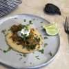 An adobo inspired chicken and kale taco with a chickpea and cassava tortilla displayed on a rustic light gray dinner plate, garnished with coconut yogurt and chopped cilantro, 3 small slices of lime are on the plate and a striped white and grey napkin, a dark rustic fork and an ancho chili are in the background