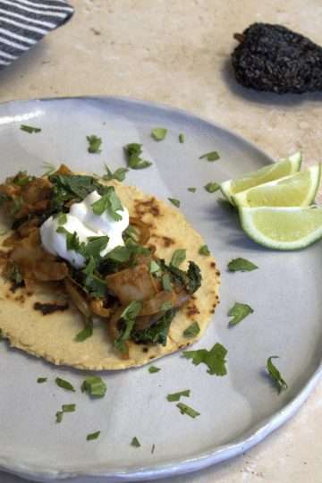 An adobo inspired chicken and kale taco with a chickpea and cassava tortilla displayed on a rustic light gray dinner plate, garnished with coconut yogurt and chopped cilantro, 3 small slices of lime are on the plate and a striped white and grey napkin, a dark rustic fork and an ancho chili are in the background
