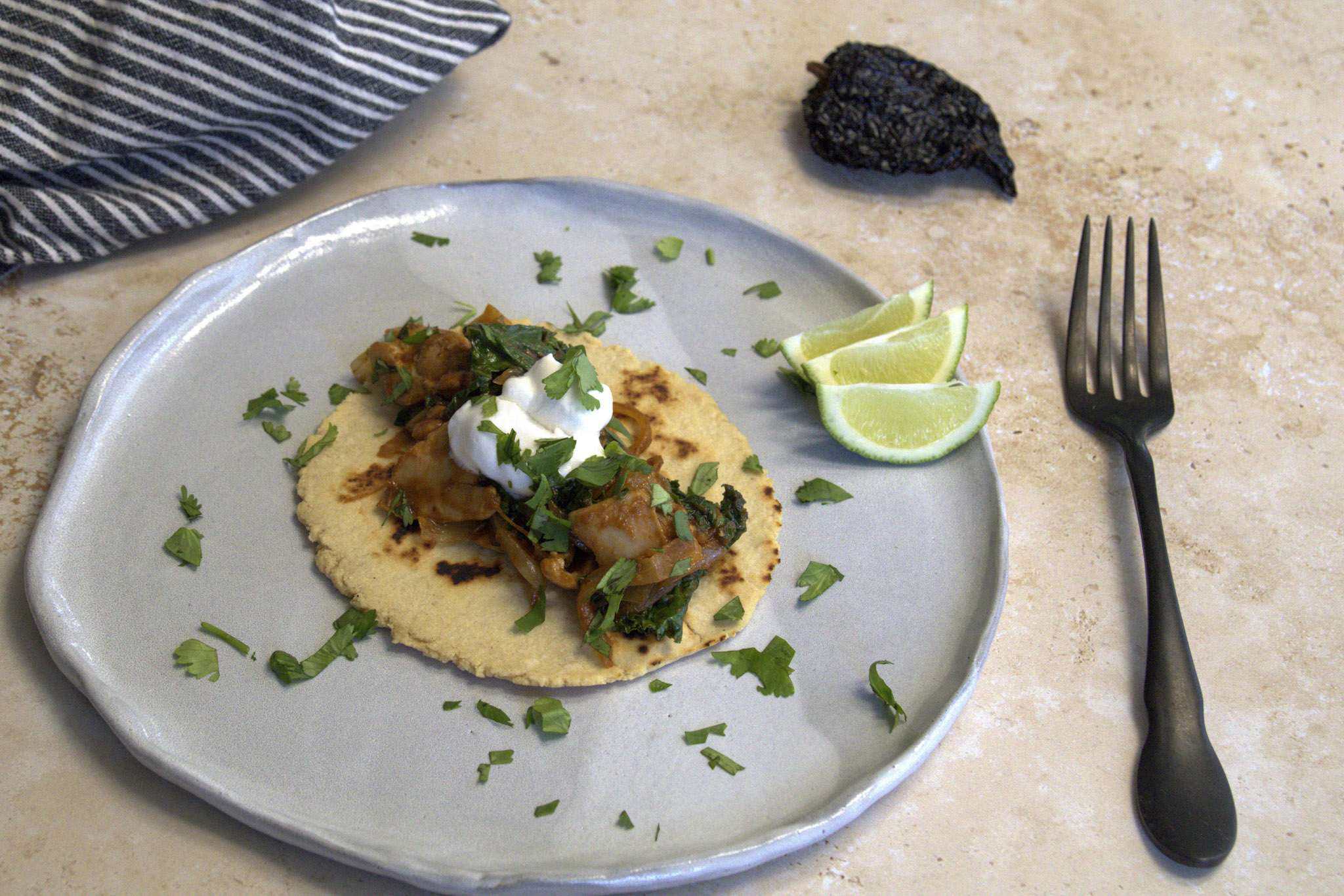 An adobo inspired chicken and kale taco with a chickpea and cassava tortilla displayed on a rustic light gray dinner plate, garnished with coconut yogurt and chopped cilantro, 3 small slices of lime are on the plate and a striped white and grey napkin, a dark rustic fork and an ancho chili are in the background