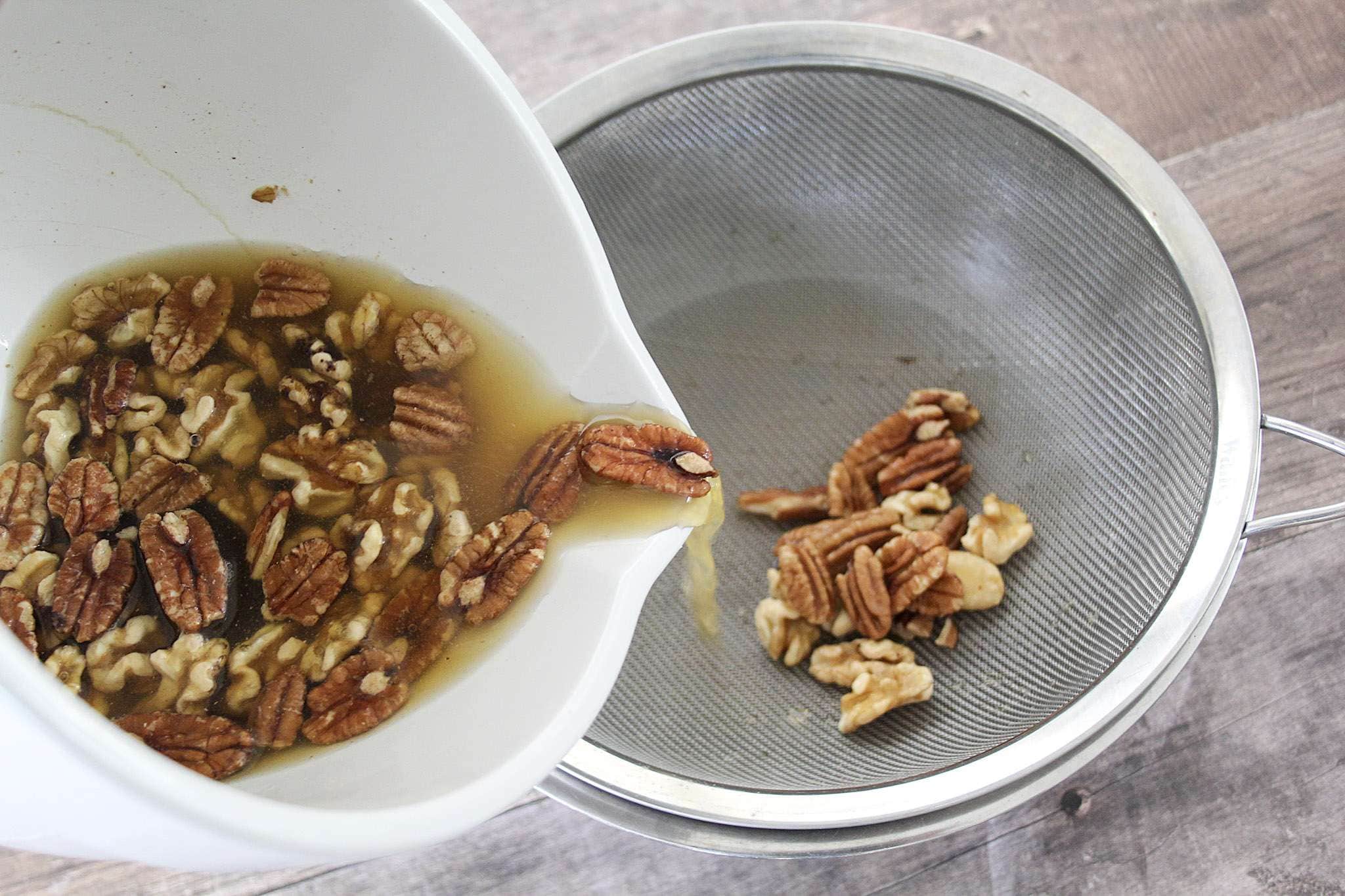 A white bowl with a pouring spout is at an angle and nits along with soaking water are being poured out through a fine mesh sieve and into another bowl