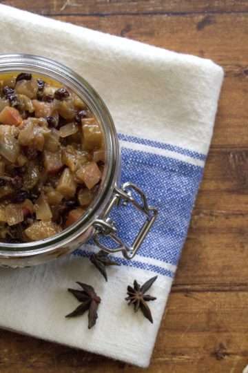 a glass jar with rhubarb chutney is resting on a white and blue kitchen towel, which is resting on a wooden surface - a piece of ginger and 3 cinnamon sticks are in the picture and a couple pieces of star anise are resting on the kitchen towel