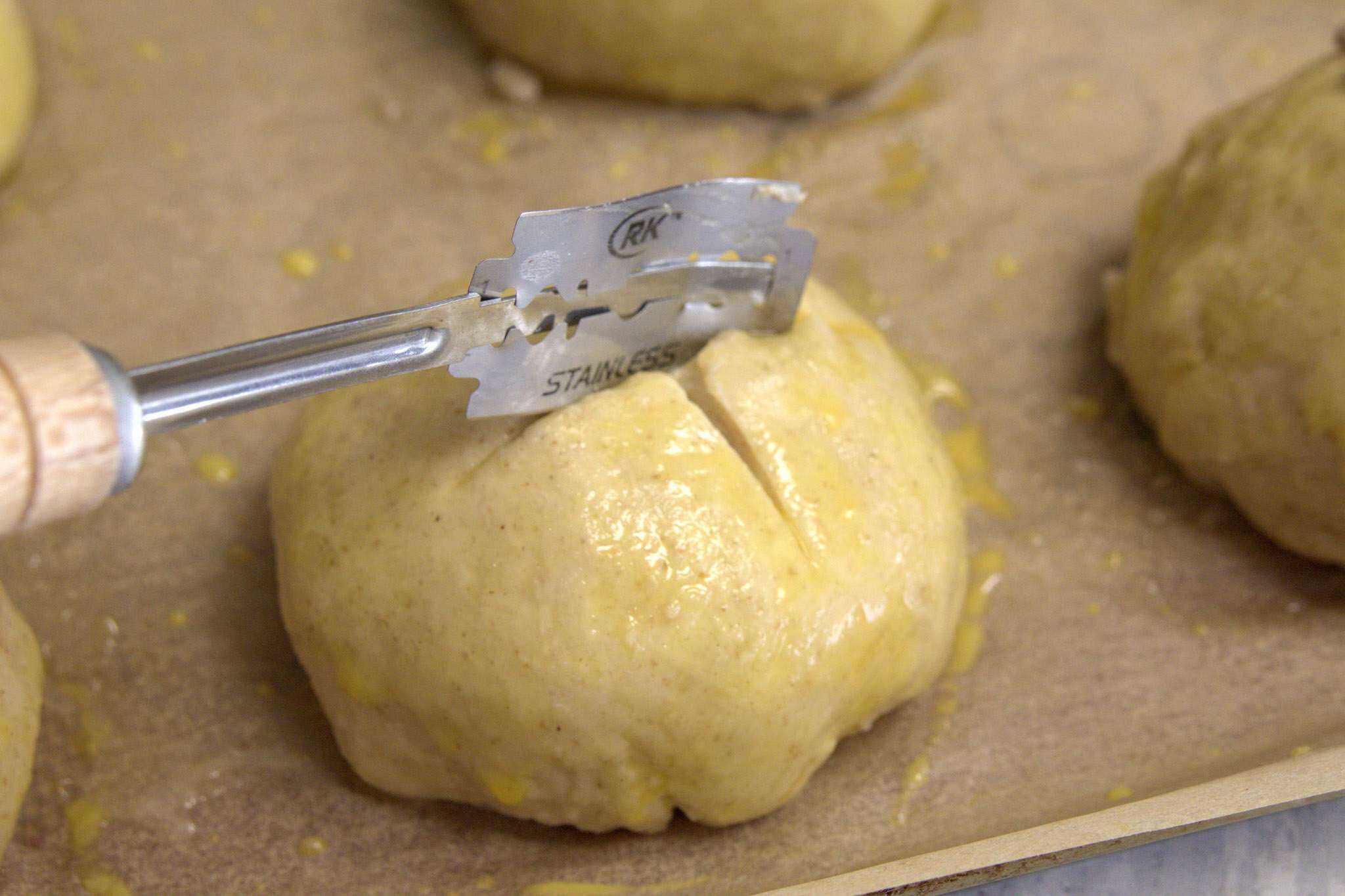 A previously boiled sourdough pretzel roll, which has been brushed with egg wash is on a parchment lined baking sheet and is being scored with a bread lame