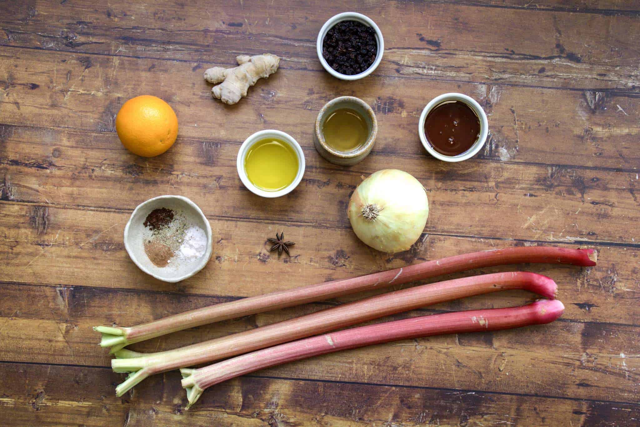 All ingredients for rhubarb chutney are displayed on a wooden surface, including rhubarb stalks, an onion, an orange, ginger, zante currants, maple syrup, apple cider vinegar, extra virgin olive oil, salt, pepper, ground cloves and cinnamon