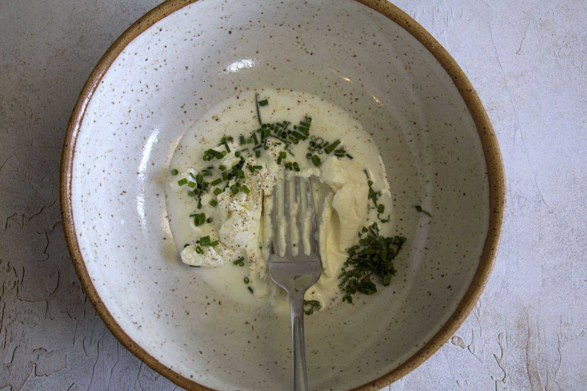 ingredient for herby goat cheese cream are in a pottery bowl with a fork resting in it