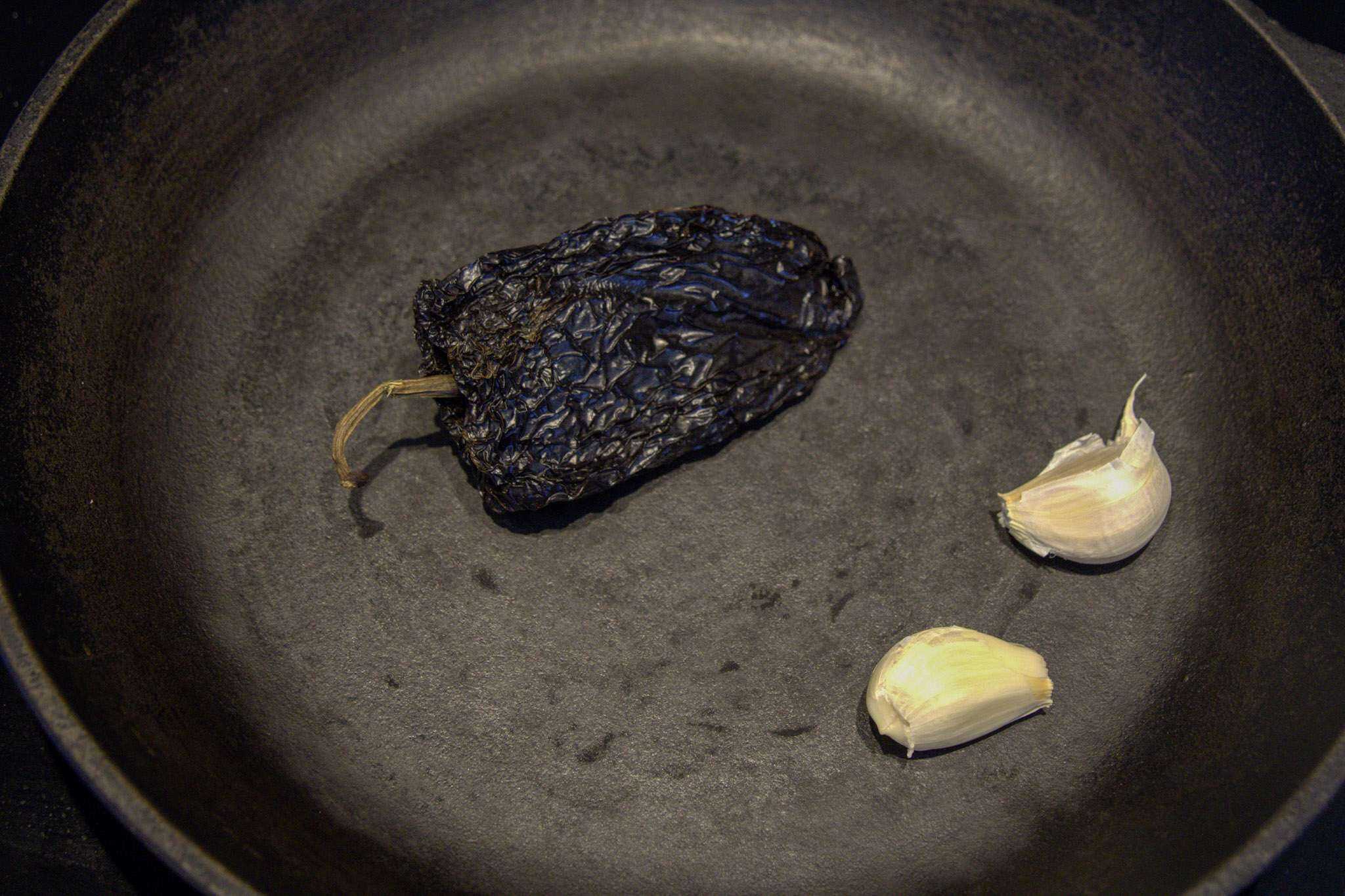 An ancho chili and two garlic cloves toasting in a cast iron pan