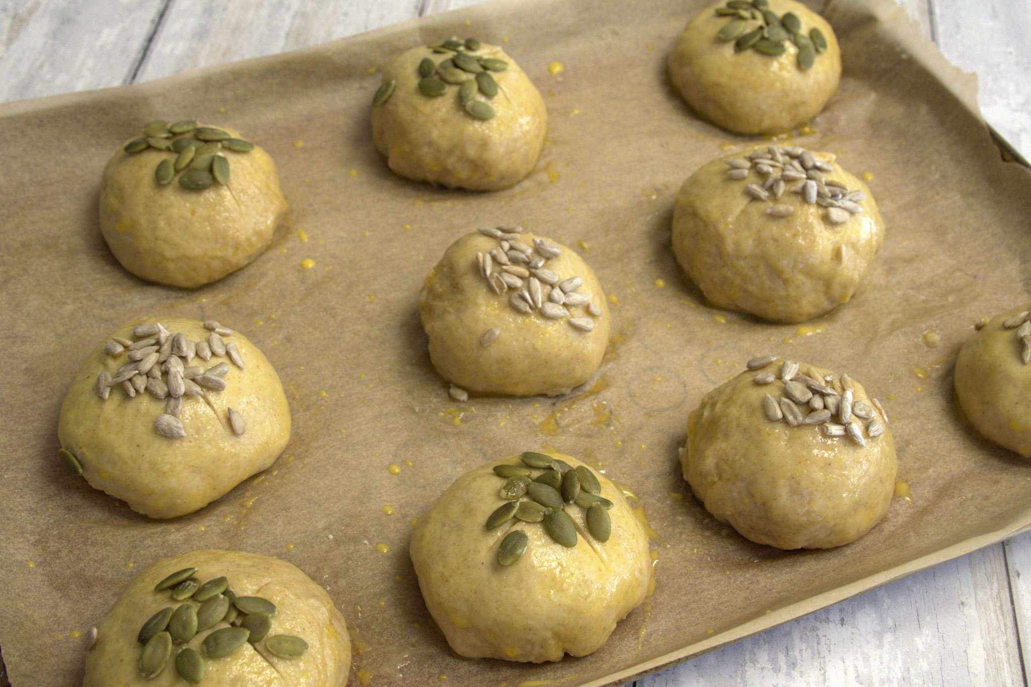 Unbaked sourdough pretzel rolls which have been brushed with egg wash, scored with a bread lame and topped with sunflower and pumpkin seeds are on a parchment lined baking sheet ready to go into the oven