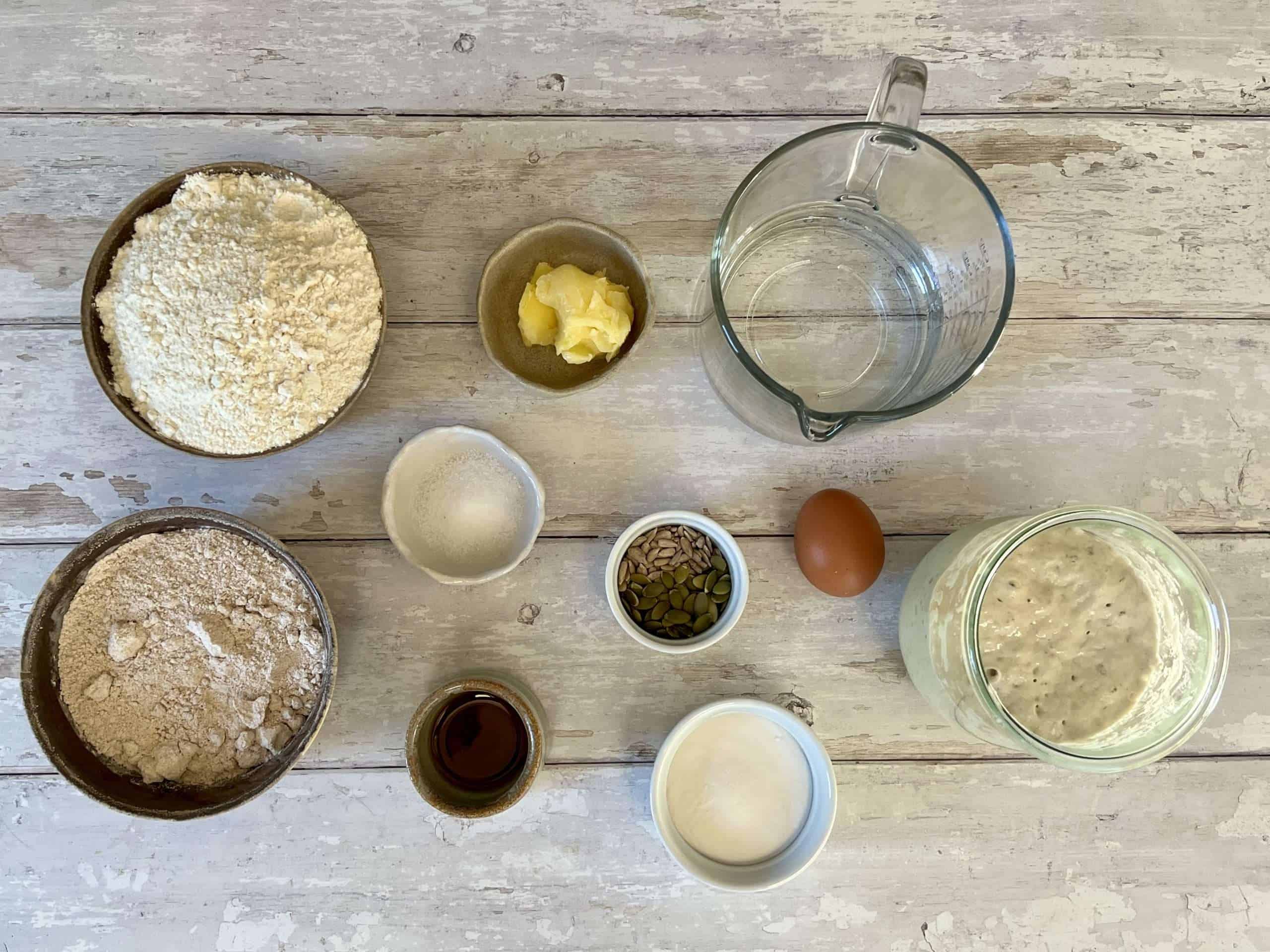 All ingredients for sourdough pretzel rolls are displayed, including bubbly sourdough starter, water, ghee, maple syrup, fine sea salt, whole spelt flour, all purpose flour, baking soda, egg, sunflower seeds and pumpkin seeds