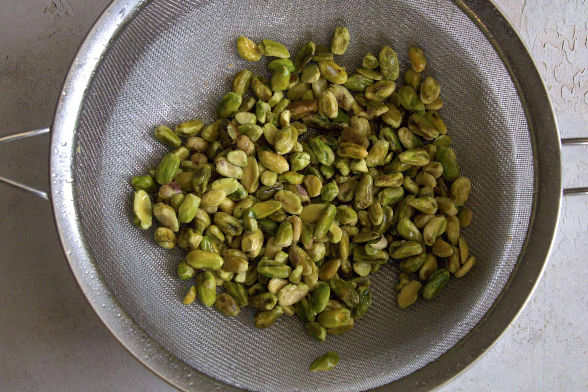 A strainer, holding just drained and rinsed soaked pistachios is shown on top of a white bowl