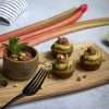 A rustic white background with a rustic wooden board in the center, holding 3 gluten-free rhubarb pistachio financiers, topped with a dollop of pistachio lime cream and a little bit of refined sugar free rhubarb compote with ginger and lime - a black fork is resting on the board and a small bowl with additional rhubarb compote was also placed on the wooden board, which is sprinkled with chopped pistachios. A few rhubarb stalks, a white and grey stiped napkin and half a lime are resting in the background.