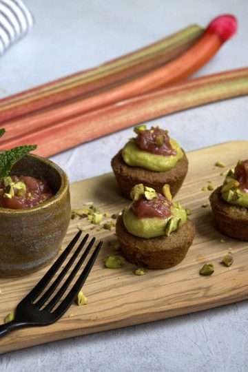 A rustic white background with a rustic wooden board in the center, holding 3 gluten-free rhubarb pistachio financiers, topped with a dollop of pistachio lime cream and a little bit of refined sugar free rhubarb compote with ginger and lime - a black fork is resting on the board and a small bowl with additional rhubarb compote was also placed on the wooden board, which is sprinkled with chopped pistachios. A few rhubarb stalks, a white and grey stiped napkin and half a lime are resting in the background.