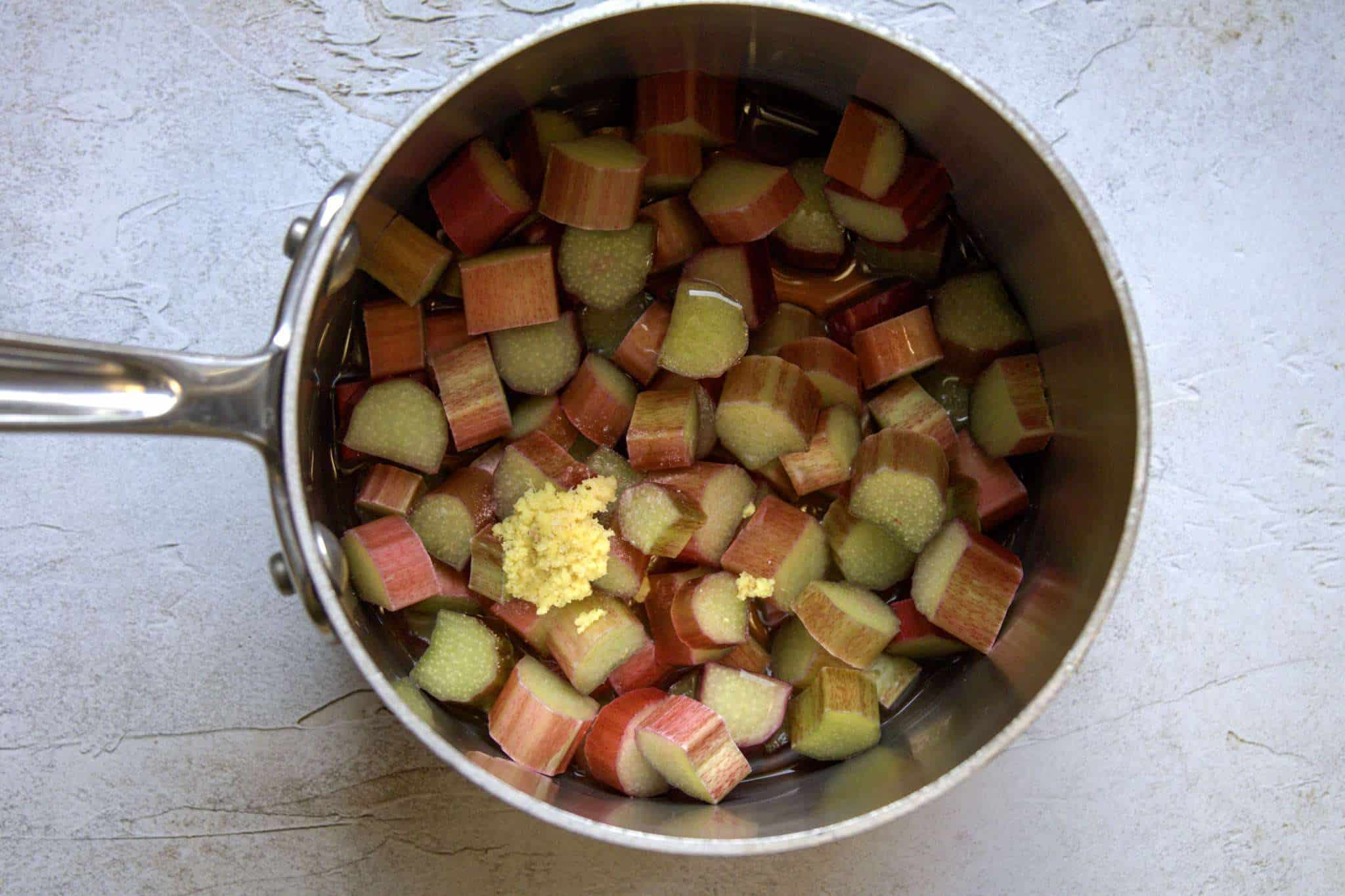 A small pot holding chopped rhubarb, along with the remaining ingredients for refined sugar-free rhubarb compote, including lime juice, grated ginger and maple syrup