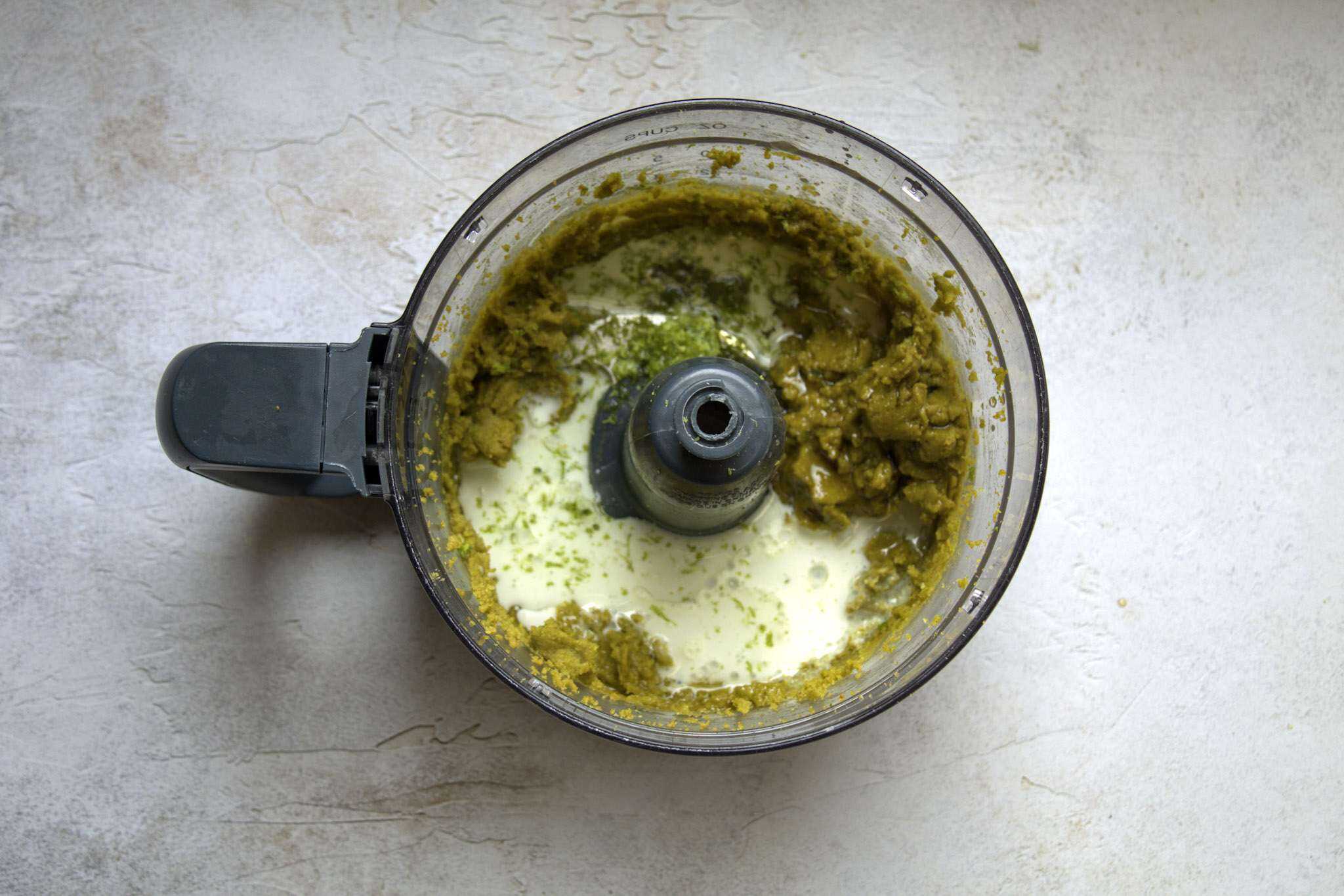 A food processor bowl is shown, holding processed pistachios with the remaining ingredients for dairy-free pistachio lime cream having just been added