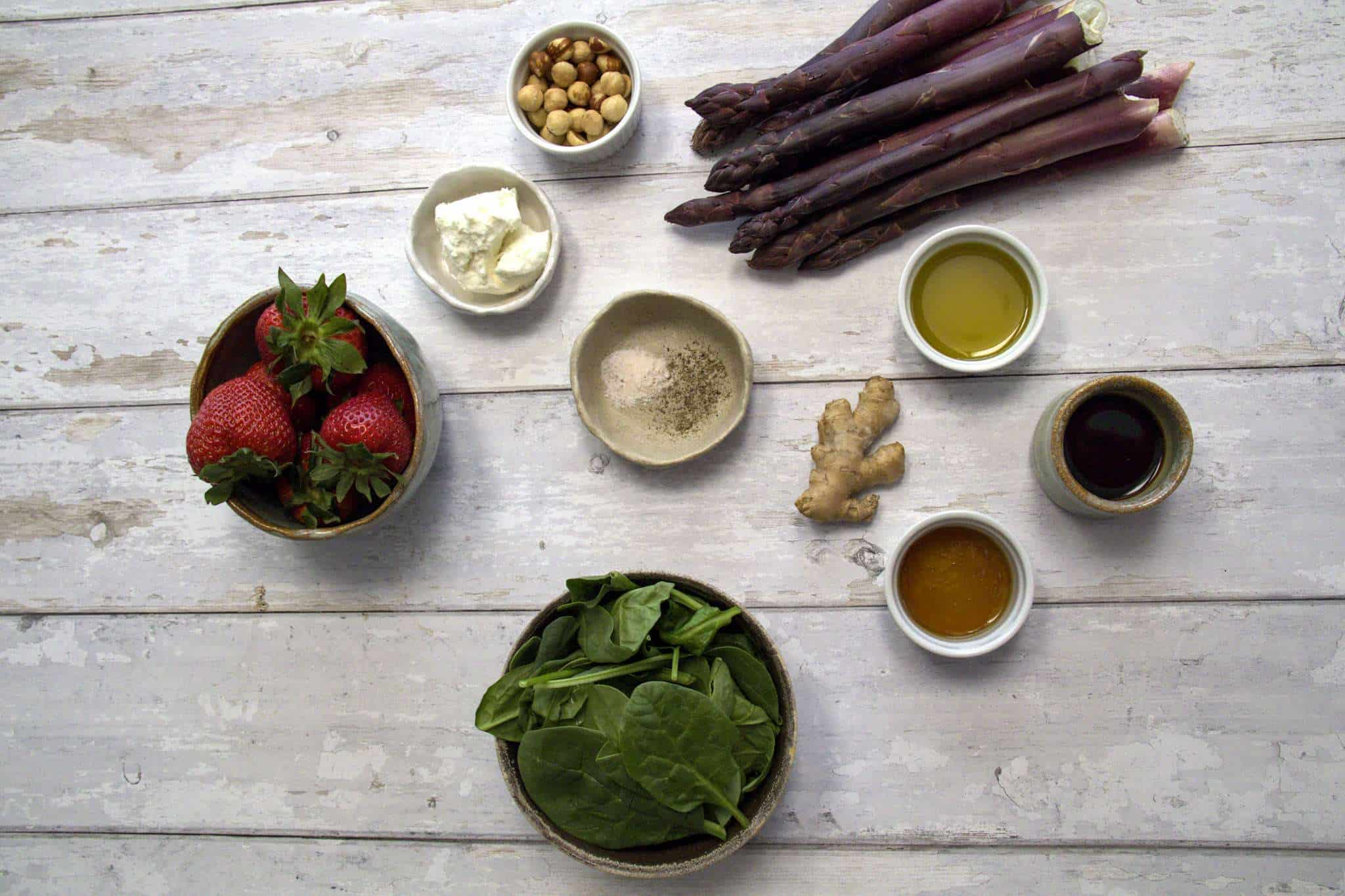 all ingredients for strawberry asparagus salad with ginger balsamic dressing are displayed on a white wooden surface, including purple asparagus spears, extra virgin olive oil, fine sea salt, black pepper, strawberries, spinach, balsamic vinegar, maple syrup, ginger, hazelnuts and goat cheese