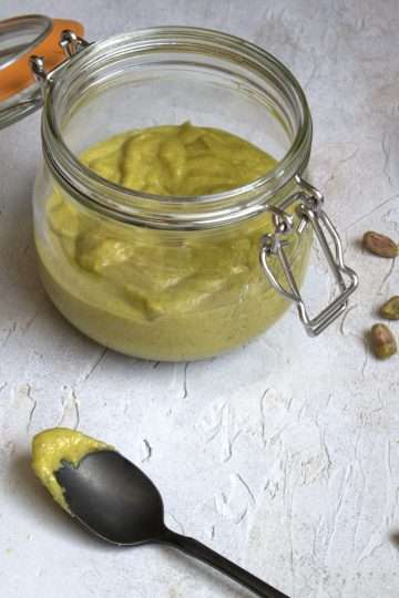 An open glass top jar holding dairy-free pistachio lime cream, in the forefront is a small pistachio rhubarb financier topped with pistachio lime cream - a black spoon is resting on the side with a little bit of pistachio lime cream on it and some whole pistachios and a whole lime are in the background