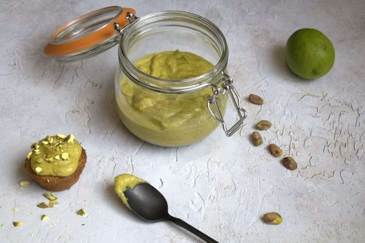 An open glass top jar holding dairy-free pistachio lime cream, in the forefront is a small pistachio rhubarb financier topped with pistachio lime cream - a black spoon is resting on the side with a little bit of pistachio lime cream on it and some whole pistachios and a whole lime are in the background