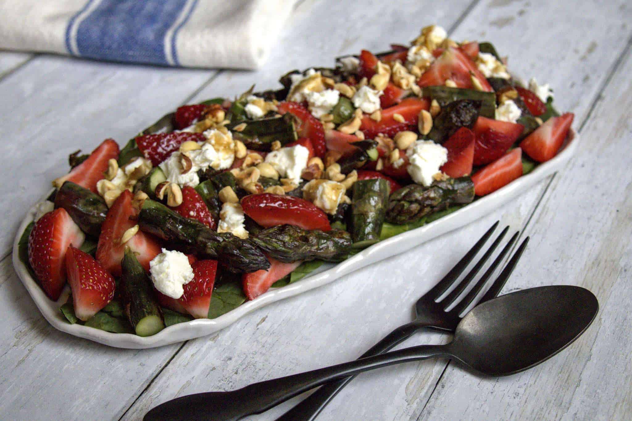 A rectangular rustic white serving platter with strawberry asparagus salad with ginger balsamic dressing is displayed in the center; a black spoon and fork in the front and a white and blue napkin is in the back - all items are displayed on a white wooden surface