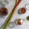 Two small open jars are displayed on the right of two large stalks of rhubarb - one is holding refined sugar-free rhubarb compote and the other contains coconut yogurt with a spoonful of rhubarb compote and a sprinkle of nourishing granola - there is also a lime in the right side of the picture. On the left side of the two rhubarb stalks is a larger jar containing rhubarb compote with a black spoon resting in it