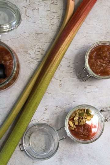 Two small open jars are displayed on the right of two large stalks of rhubarb - one is holding refined sugar-free rhubarb compote and the other contains coconut yogurt with a spoonful of rhubarb compote and a sprinkle of nourishing granola - there is also a lime in the right side of the picture. On the left side of the two rhubarb stalks is a larger jar containing rhubarb compote with a black spoon resting in it