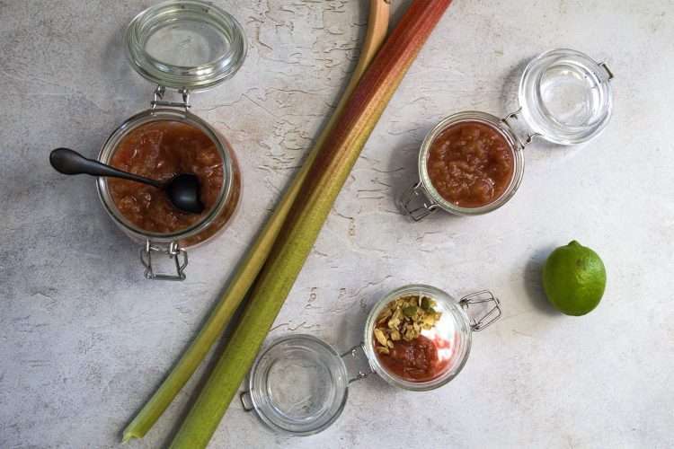 Two small open jars are displayed on the right of two large stalks of rhubarb - one is holding refined sugar-free rhubarb compote and the other contains coconut yogurt with a spoonful of rhubarb compote and a sprinkle of nourishing granola - there is also a lime in the right side of the picture. On the left side of the two rhubarb stalks is a larger jar containing rhubarb compote with a black spoon resting in it