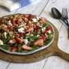 A round rustic white serving platter with strawberry asparagus salad with ginger balsamic dressing is displayed in the center of the picture; the plate is sitting on a round wooden paddle, a black spoon and fork are to the right of it and a white and blue napkin is in the back - all items are displayed on a white wooden surface