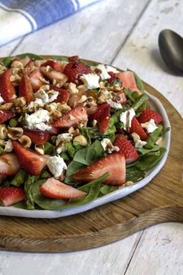 A round rustic white serving platter with strawberry asparagus salad with ginger balsamic dressing is displayed in the center of the picture; the plate is sitting on a round wooden paddle, a black spoon and fork are to the right of it and a white and blue napkin is in the back - all items are displayed on a white wooden surface